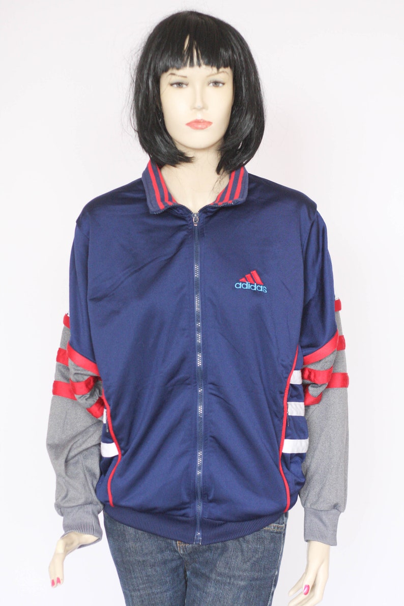 Peut inclure: Une veste zipp&eacute;e bleu marine avec des rayures rouges et blanches sur les manches. La veste a un panneau gris sous les bras et le logo Adidas sur la poitrine.