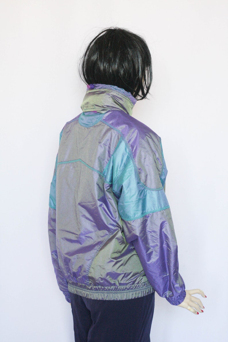 Pu&ograve; includere: Una giacca iridescente viola e turchese con chiusura a zip. La giacca ha un colletto alto e maniche lunghe.