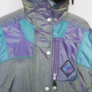 Pu&ograve; includere: Una giacca blu e viola iridescente con una tasca con cerniera e un logo sul davanti. La giacca ha un colletto alto e una chiusura a scatto.