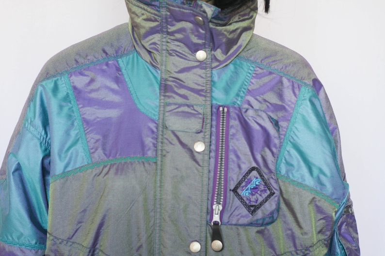 Pu&ograve; includere: Una giacca blu e viola iridescente con una tasca con cerniera e un logo sul davanti. La giacca ha un colletto alto e una chiusura a scatto.