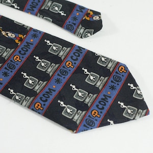 May include: A navy blue tie featuring a repeating pattern of computer monitors, exclamation marks, and the text ".COM *!@?". The design incorporates blue and orange stripes. The tie is made of a smooth fabric.
