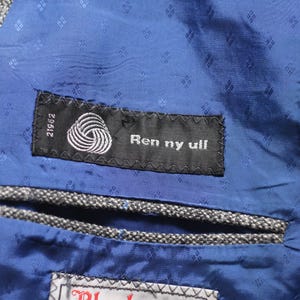 Pu&ograve; includere: Un primo piano di una fodera in tessuto blu con un'etichetta nera che recita "Ren ny ull" e un simbolo di lana bianco. L'etichetta &egrave; attaccata a un tessuto in tweed grigio.