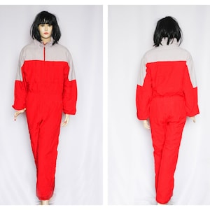 Peut inclure: Combinaison de ski rouge et grise avec une fermeture éclair. La combinaison a un col montant et des manches longues.