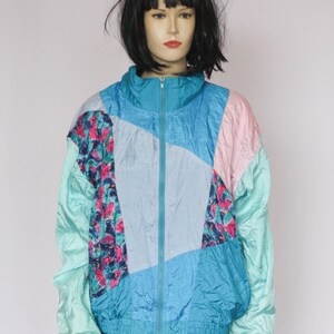 Puede incluir: Una chaqueta de color azul, rosa y verde con bloques de color y cierre de cremallera. La chaqueta tiene un patr&oacute;n geom&eacute;trico y est&aacute; hecha de un material brillante similar al nailon.
