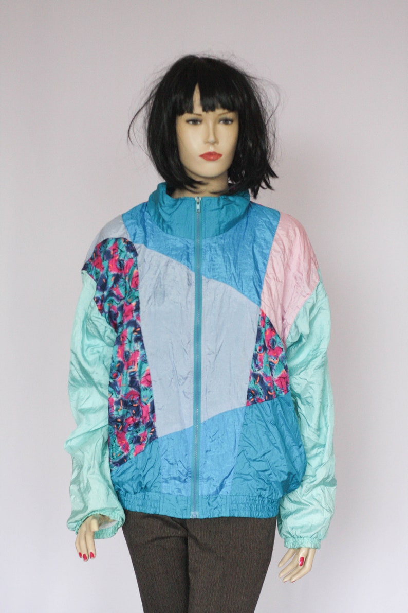 Puede incluir: Una chaqueta de color azul, rosa y verde con bloques de color y cierre de cremallera. La chaqueta tiene un patr&oacute;n geom&eacute;trico y est&aacute; hecha de un material brillante similar al nailon.