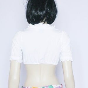 Pu&ograve; includere: Blusa bianca corta con maniche corte e scollo a volant. La blusa &egrave; indossata sopra una gonna a stampa floreale con fiori rosa, gialli e verdi.