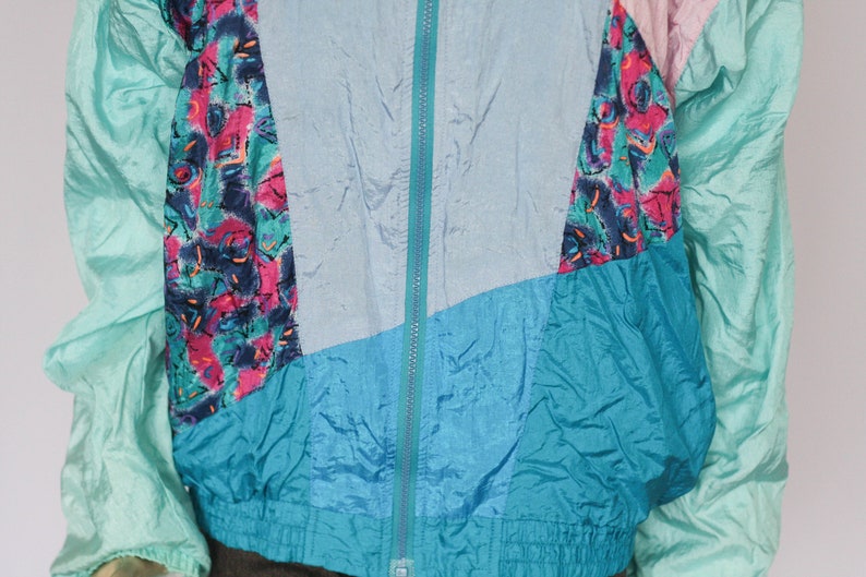 Puede incluir: Una chaqueta vintage multicolor con cierre de cremallera. La chaqueta est&aacute; hecha de un material brillante similar al nailon y presenta un patr&oacute;n geom&eacute;trico en tonos de azul, verde, rosa y blanco.