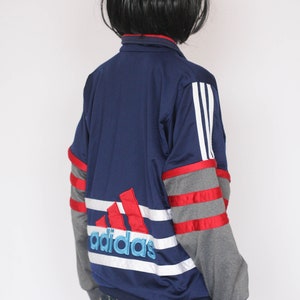 Peut inclure: Une veste de surv&ecirc;tement Adidas bleu marine avec des rayures rouges, blanches et grises. Le logo Adidas est brod&eacute; en rouge, blanc et bleu au dos.