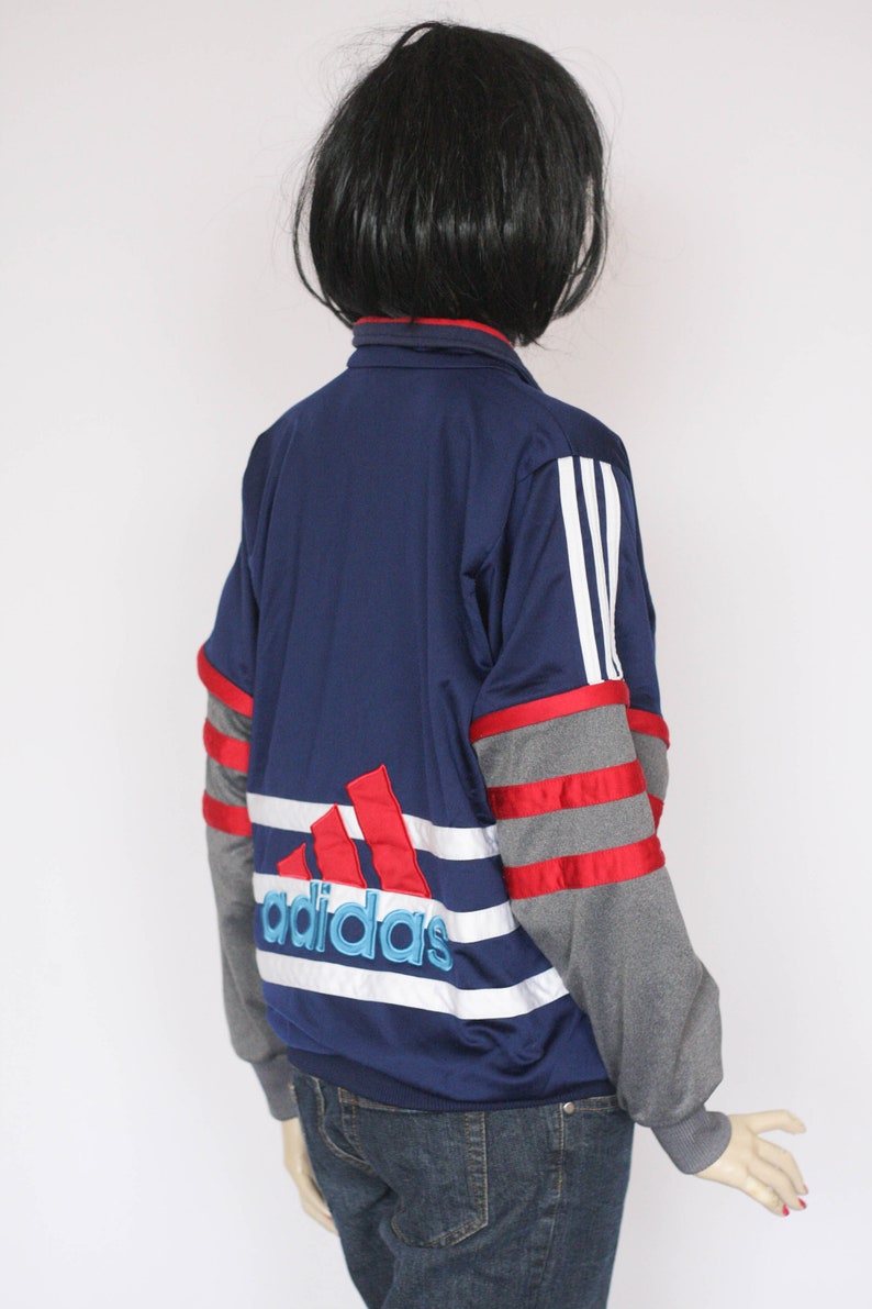 Peut inclure: Une veste de surv&ecirc;tement Adidas bleu marine avec des rayures rouges, blanches et grises. Le logo Adidas est brod&eacute; en rouge, blanc et bleu au dos.