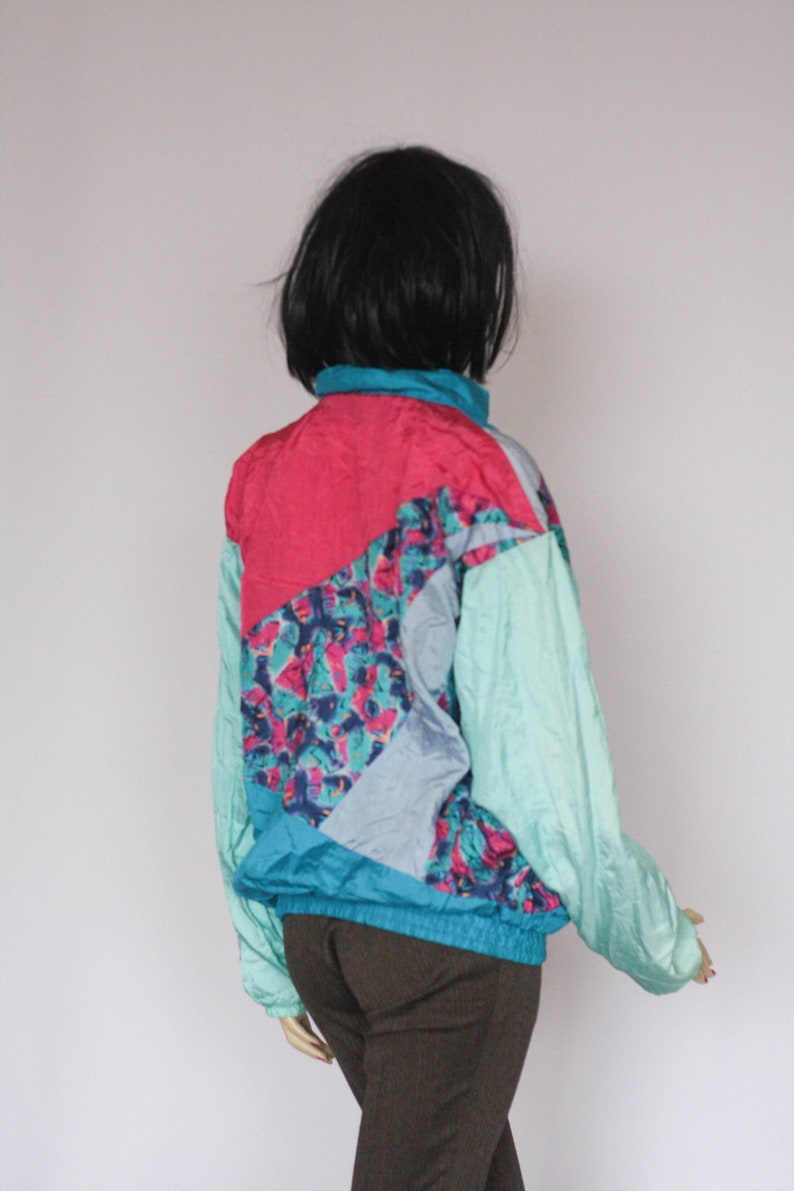 Puede incluir: Una chaqueta cortavientos azul, rosa y turquesa con un patr&oacute;n geom&eacute;trico. La chaqueta tiene una cremallera y un cuello alto.