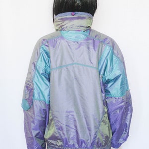 Pu&ograve; includere: Una giacca iridescente viola e turchese con chiusura a zip. La giacca ha un colletto alto e una finitura riflettente.