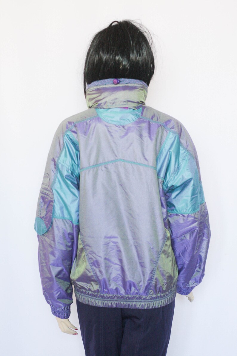 Pu&ograve; includere: Una giacca iridescente viola e turchese con chiusura a zip. La giacca ha un colletto alto e una finitura riflettente.