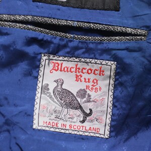 Pu&ograve; includere: Una tasca in tessuto blu con un'etichetta che recita "Blackcock Rug Reg'd Made in Scotland". L'etichetta presenta un'illustrazione in bianco e nero di un uccello.