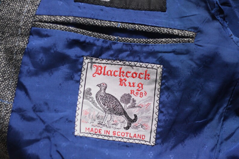 Pu&ograve; includere: Una tasca in tessuto blu con un'etichetta che recita "Blackcock Rug Reg'd Made in Scotland". L'etichetta presenta un'illustrazione in bianco e nero di un uccello.