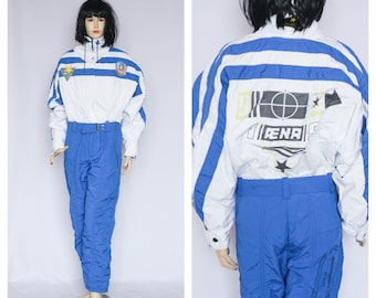 May include: White and blue ski suit with a striped pattern and multiple logos, including "AENA" on the back. The suit has a blue belt and a zipper closure.