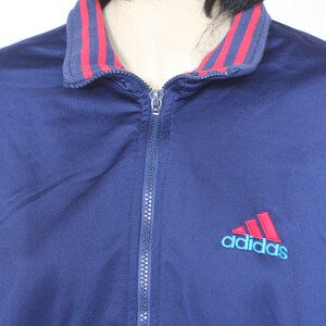 Peut inclure: Une veste zipp&eacute;e bleu marine avec un col ray&eacute; rouge et blanc et le logo Adidas en rouge et bleu.