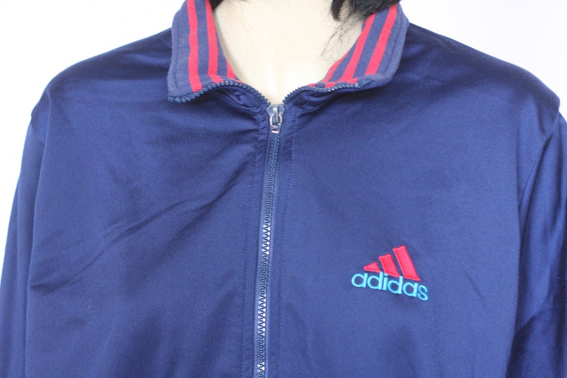 Peut inclure: Une veste zipp&eacute;e bleu marine avec un col ray&eacute; rouge et blanc et le logo Adidas en rouge et bleu.