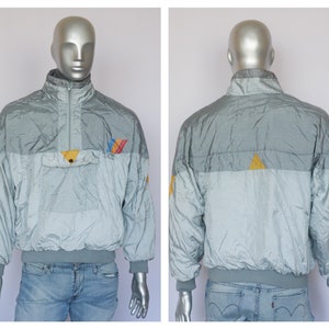 Op de afbeelding: Een zilveren, blauwe en gele windbreaker-jas met een ritssluiting en een klein zakje op de borst.