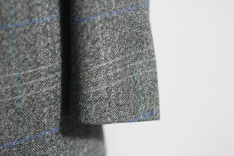 Pu&ograve; includere: Primo piano di un tessuto a quadri grigio e blu con una texture sottile. Il tessuto presenta un motivo intrecciato con sottili linee blu.