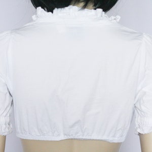 Pu&ograve; includere: Blusa bianca a maniche corte con colletto a volant e polsini elastici.