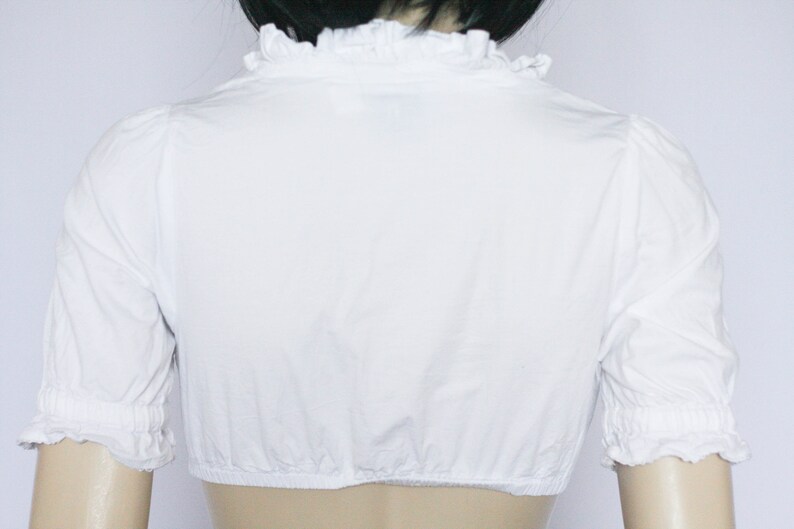 Pu&ograve; includere: Blusa bianca a maniche corte con colletto a volant e polsini elastici.