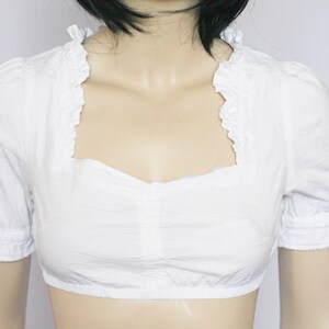 Pu&ograve; includere: Blusa bianca corta con scollo a volant e maniche corte.