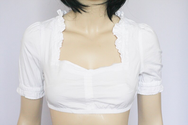 Pu&ograve; includere: Blusa bianca corta con scollo a volant e maniche corte.
