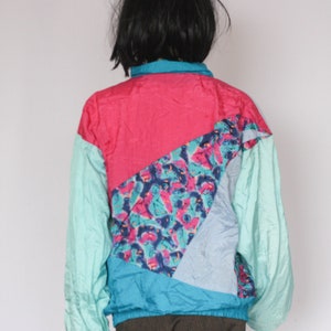 Puede incluir: Una chaqueta colorida, multicolor con un dise&ntilde;o geom&eacute;trico. La chaqueta est&aacute; hecha de una tela ligera de nylon y tiene una cremallera. La chaqueta est&aacute; en un esquema de colores turquesa, rosa, azul y blanco.