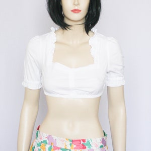 Pu&ograve; includere: Una blusa bianca corta con scollo a volant e maniche corte. La blusa &egrave; abbinata a una gonna floreale colorata.