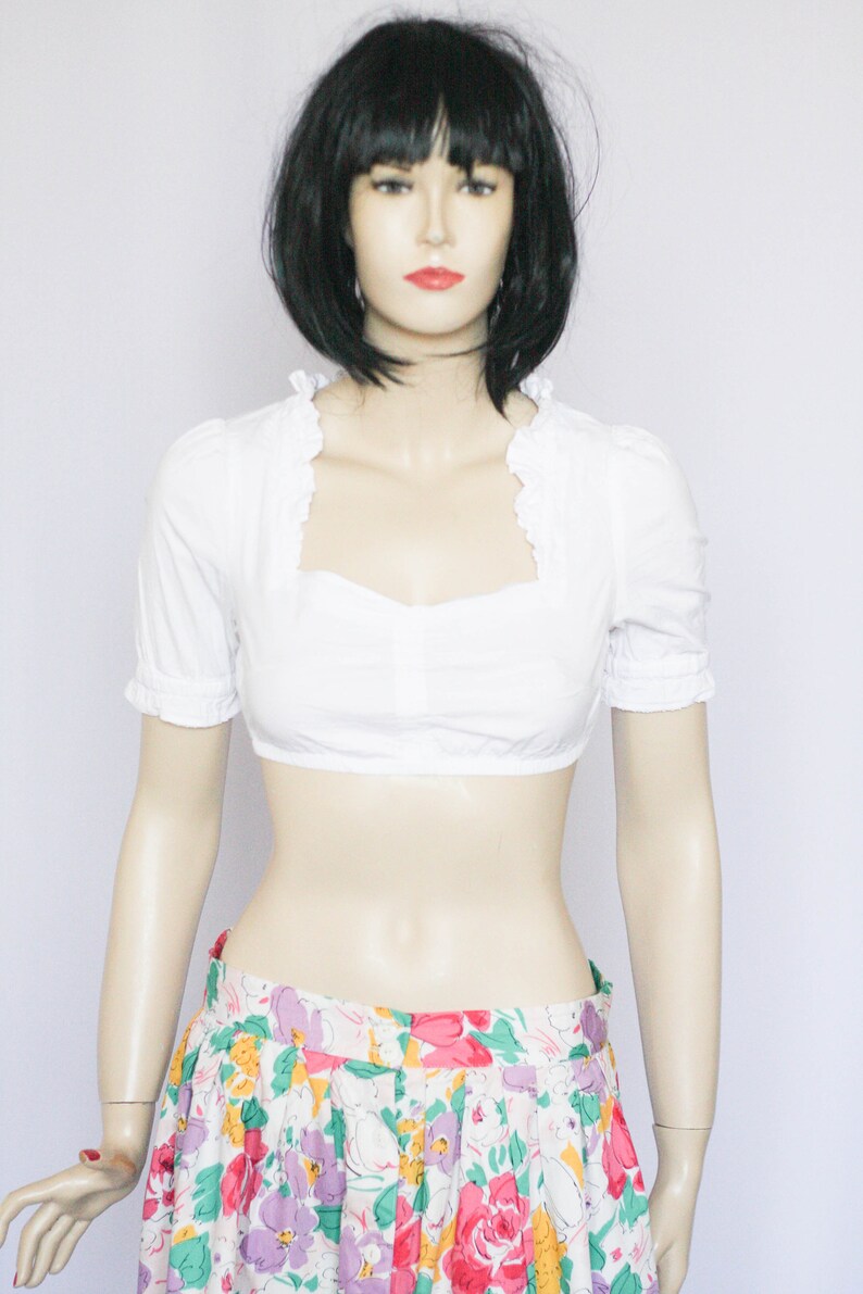 Pu&ograve; includere: Una blusa bianca corta con scollo a volant e maniche corte. La blusa &egrave; abbinata a una gonna floreale colorata.