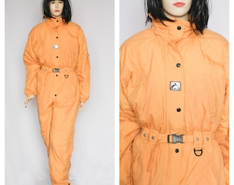 Könnte beinhalten: Orangefarbener Ski-Overall mit schwarzem Gürtel und einem Logo auf der Brust. Der Overall hat einen Reißverschluss und einen hohen Kragen.