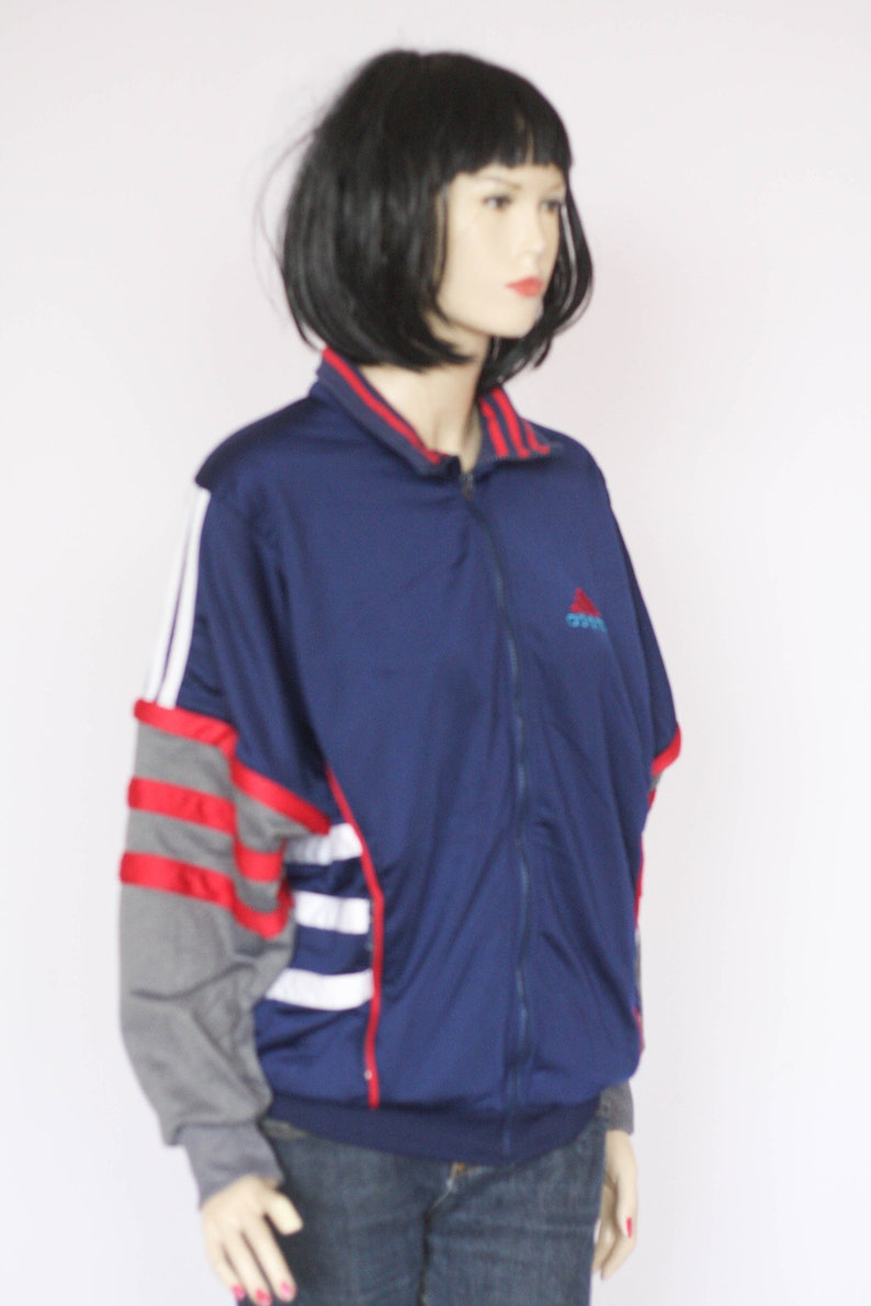 Peut inclure: Une veste zipp&eacute;e bleu marine avec des rayures rouges, blanches et grises sur les manches. La veste a un petit logo Adidas sur la poitrine.