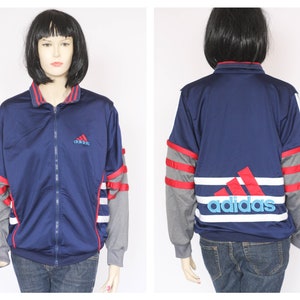 Peut inclure: Une veste zipp&eacute;e bleu marine avec des rayures rouges et blanches sur les manches et un grand logo Adidas blanc et bleu au dos. La veste a un petit logo Adidas brod&eacute; sur la poitrine gauche.