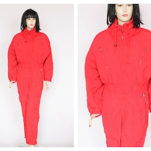 Peut inclure: Une combinaison de ski rouge avec une fermeture éclair et une capuche à cordon. La combinaison a plusieurs poches avec des fermetures éclair.