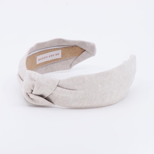 A light tan knotted headband made of linen fabric. The headband has a label that says "MADDIE AND ME".