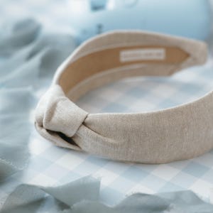 May include: A light beige knotted headband made of linen fabric. The headband is on a blue and white gingham background.
