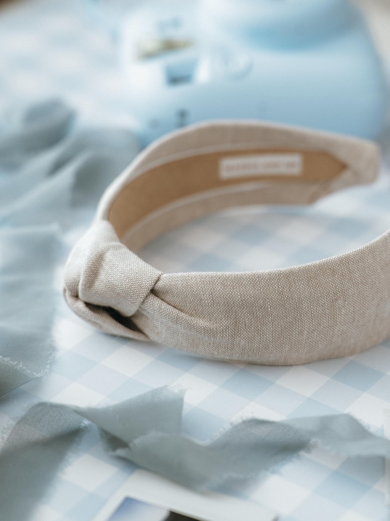 May include: A light beige knotted headband made of linen fabric. The headband is on a blue and white gingham background.