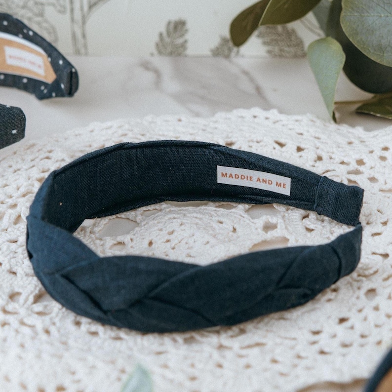 May include: A dark navy blue braided headband made of linen fabric. The headband has a label that says "MADDIE AND ME".