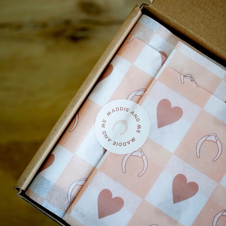 May include: A white gift box with a pink and white checkered tissue paper lining. The tissue paper has a repeating pattern of hearts and horseshoes. A white sticker with the text "MADDIE AND ME" is on the tissue paper.