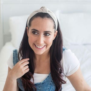 May include: A woman with long dark hair is wearing a light beige knotted headband. She is smiling and looking to the side. She is wearing a white t-shirt and a denim overall dress.
