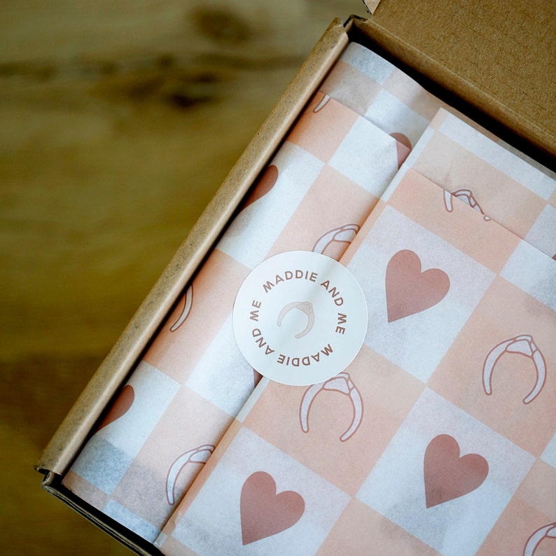 May include: A white and pink checkered gift wrapping paper with a round sticker that says "Maddie and Me" with a horseshoe design. The sticker is placed on the top left corner of the paper.