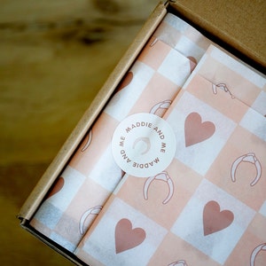 May include: A white sticker with the text "Maddie and Me" in a circle on a pink and white checkered tissue paper. The sticker has a small illustration of a headband.