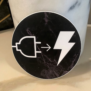 May include: A black and white sticker with a plug and lightning bolt graphic on a black marble background. The graphic shows a plug with an arrow pointing to a lightning bolt. The text "Plug in to Power" is written below the graphic.