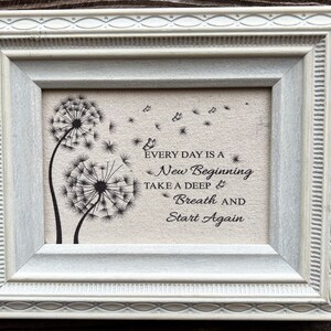 May include: A framed print with a white frame and a beige background. The print features a black and white illustration of two dandelions with seeds blowing in the wind. The text "Every day is a new beginning. Take a deep breath and start again." is printed below the dandelions.