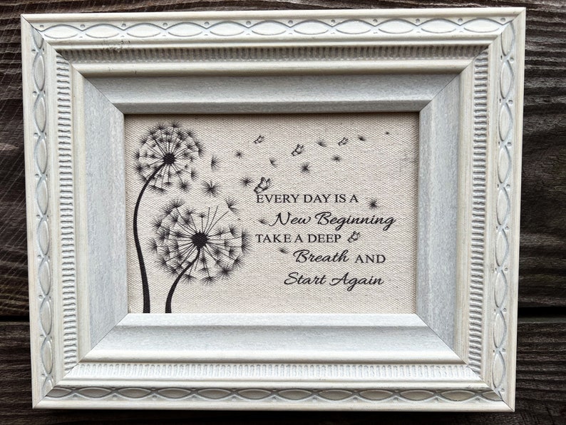 May include: A framed print with a white frame and a beige background. The print features a black and white illustration of two dandelions with seeds blowing in the wind. The text "Every day is a new beginning. Take a deep breath and start again." is printed below the dandelions.