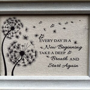 May include: Framed canvas print with a quote about new beginnings. The quote reads "Every day is a new beginning. Take a deep breath and start again." The print features two black dandelion silhouettes with seeds blowing in the wind.