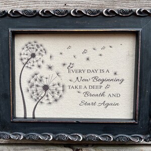 May include: A framed print with a burlap background featuring two dandelion illustrations and the text "Every day is a new beginning. Take a deep breath and start again."