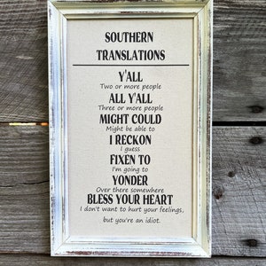 May include: A white framed sign with a beige background and black text. The sign reads "Southern Translations" and lists common Southern sayings and their meanings. For example, "Y'all" is defined as "Two or more people" and "All Y'all" is defined as "Three or more people".