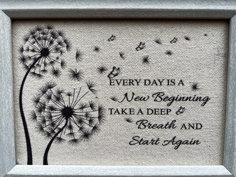 May include: A framed canvas print with a quote about new beginnings. The quote reads "Every day is a new beginning. Take a deep breath and start again." The print features two black dandelion silhouettes with seeds blowing in the wind.