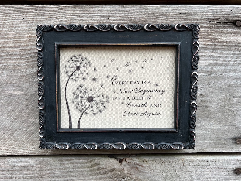 May include: Black framed canvas print with a quote about new beginnings. The quote reads "Every day is a new beginning. Take a deep breath and start again." The print features two dandelion illustrations with seeds blowing in the wind.